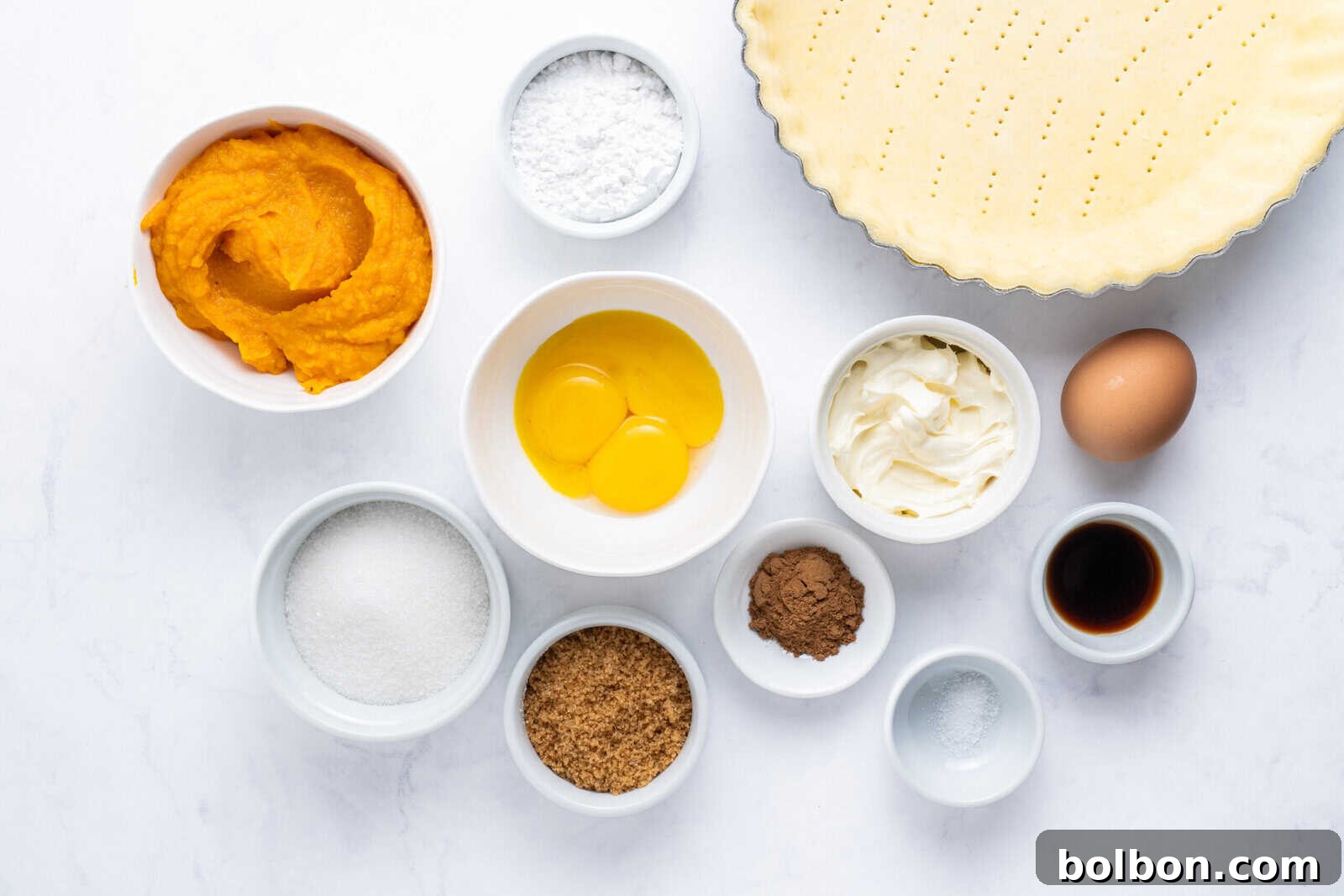A flat lay of fresh pumpkin pie ingredients including pumpkin purée, eggs, sugars, cocoa powder, salt, vanilla extract, cream cheese, powdered sugar, and an unbaked pie crust.