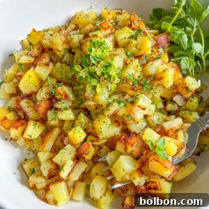 A white bowl filled with golden, diced roasted potatoes garnished with fresh green herbs, with a fork and a small bunch of leafy greens on the side.
