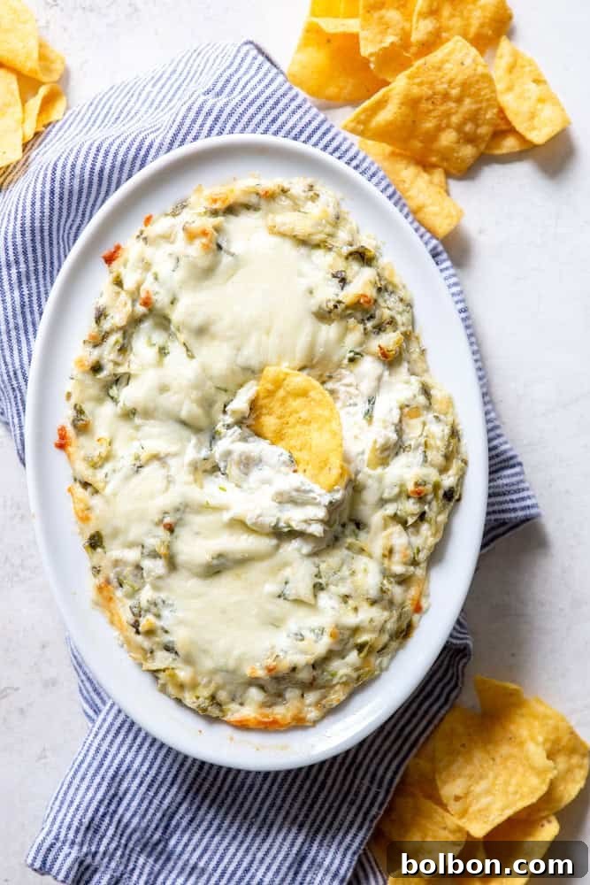 Close-up of creamy, hot Spinach Artichoke Dip in a white dish, garnished with fresh parsley, sitting on a blue and white striped dishcloth.