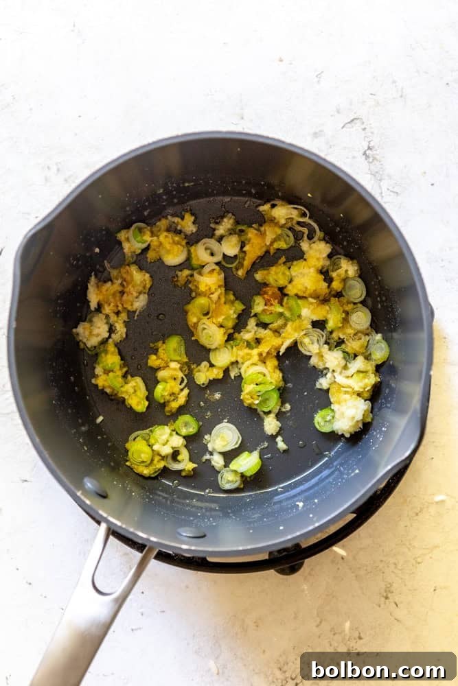 Fresh ginger, garlic, and the white parts of scallions sautéing in avocado and sesame oil in a pan, releasing their fragrant aromas.