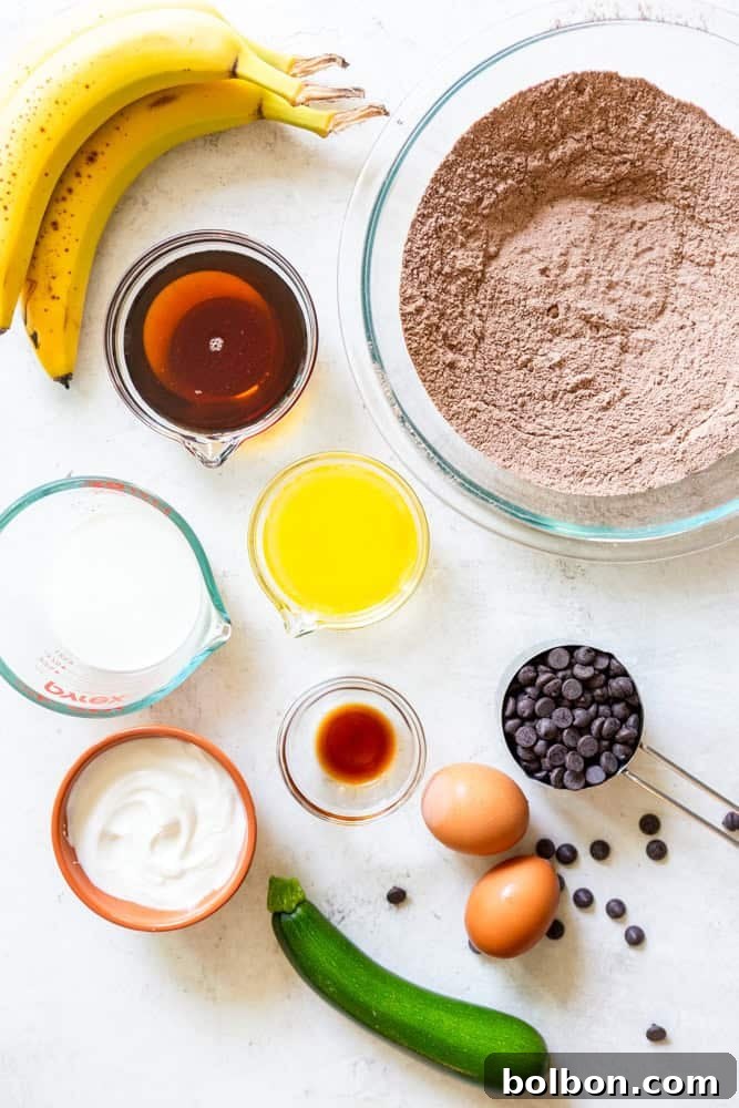 All the wholesome ingredients for healthy chocolate zucchini muffins: ripe bananas, maple syrup, melted butter, milk, Greek yogurt, vanilla extract, shredded zucchini, eggs, chocolate chips, golden whole wheat flour, cocoa powder, baking powder, baking soda, salt, and a hint of cinnamon.