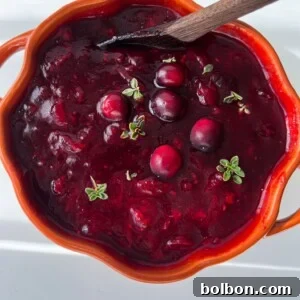 A bright orange bowl filled with thick cranberry sauce, garnished with whole cranberries and small green herb sprigs.