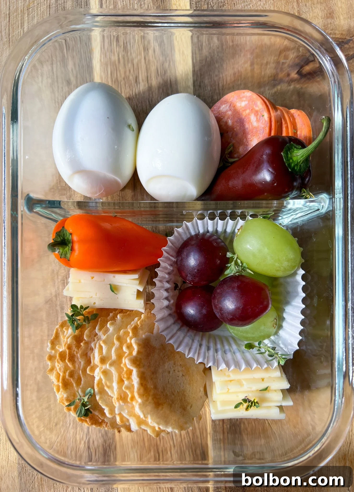 A beautifully arranged Adult Lunchable featuring hard-boiled eggs, pepperoni, cubed Havarti cheese, grapes, and crisps in a segmented glass meal prep container, ready for a nutritious and convenient meal.