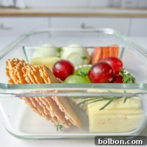 A glass container filled with sliced crackers, cheese, green and red grapes, carrot sticks, and cucumber slices creates the perfect Adult Lunchable, placed on a white surface with a blurred kitchen background.