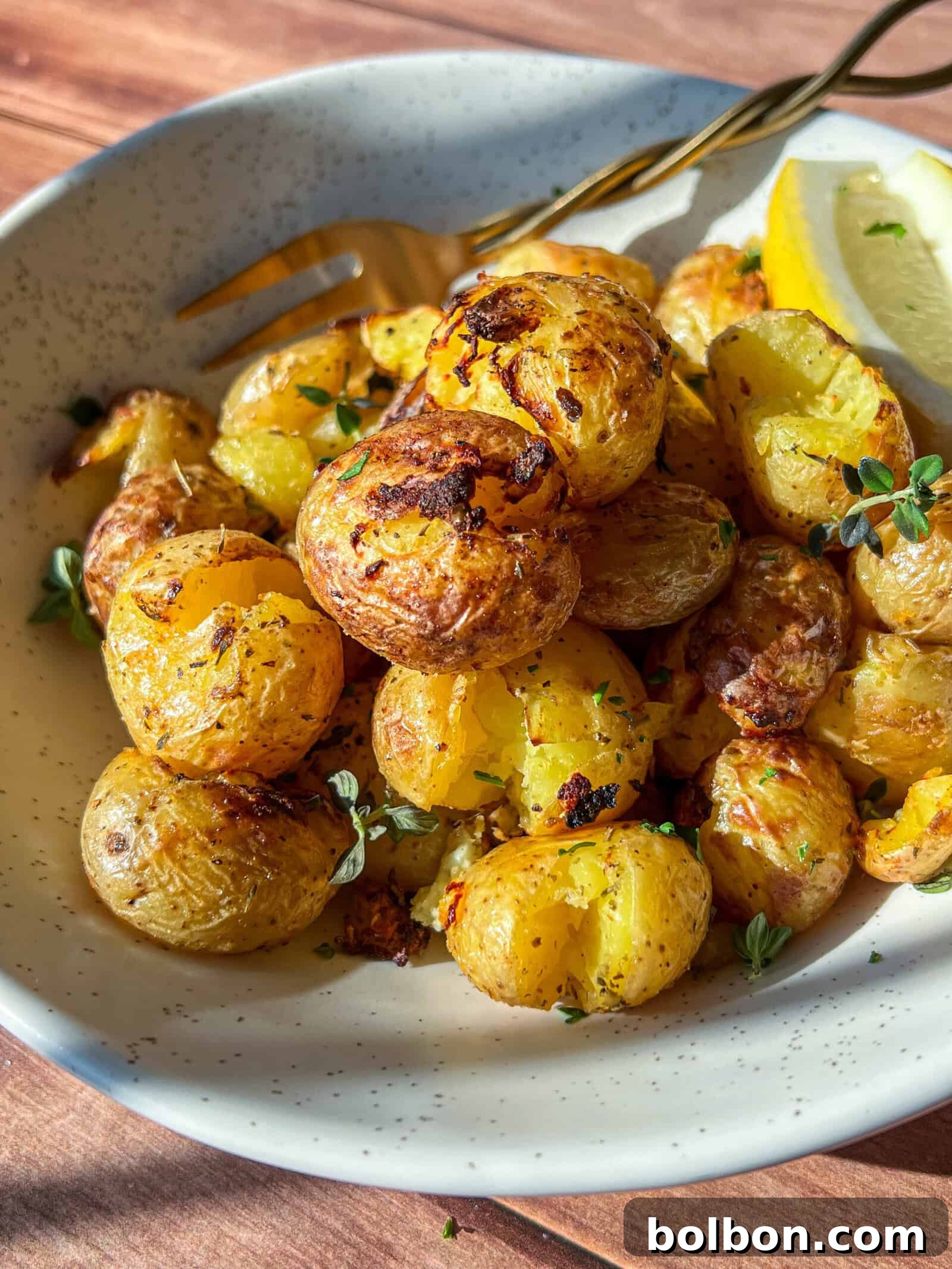 A bowl of crispy roasted baby potatoes garnished with fresh herbs, served with a lemon wedge and a fork on the side, all on a speckled ceramic ceramic plate.