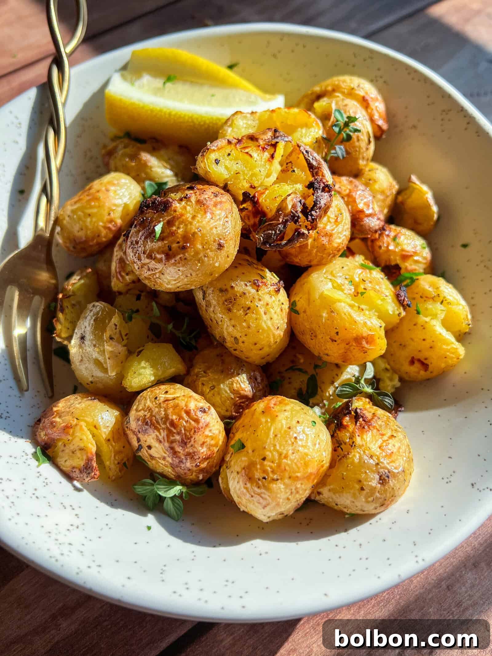 A white speckled bowl filled with roasted baby potatoes garnished with herbs and two lemon wedges, with a gold fork resting on the edge, on a wooden surface.
