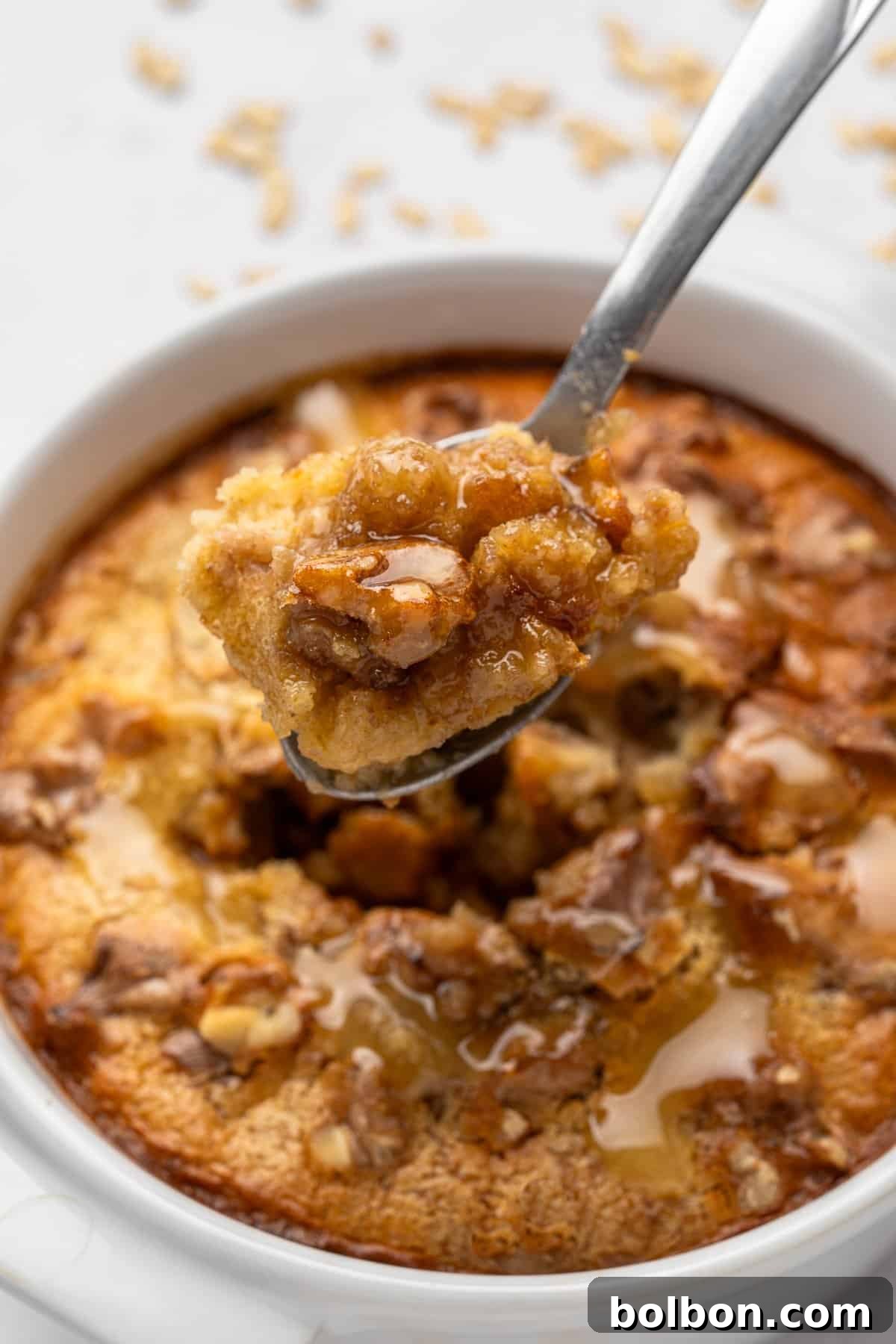 A spoonful of warm bread pudding with walnuts and honey, held above a white ramekin filled with more pudding. The dessert has a golden-brown crust and is drizzled with honey, showing a moist, flavorful texture.