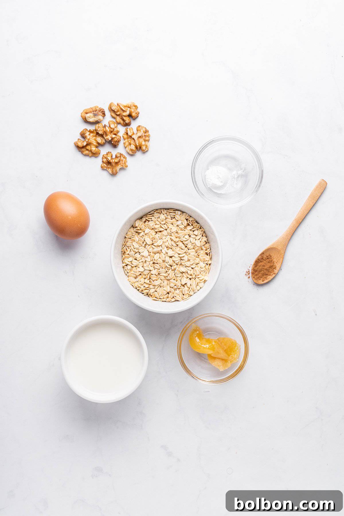 Ingredients for a recipe laid out on a white surface: a bowl of oats, a small bowl of milk, an egg, walnut pieces, a spoonful of cinnamon, a bowl of honey, and a small dish of baking soda.