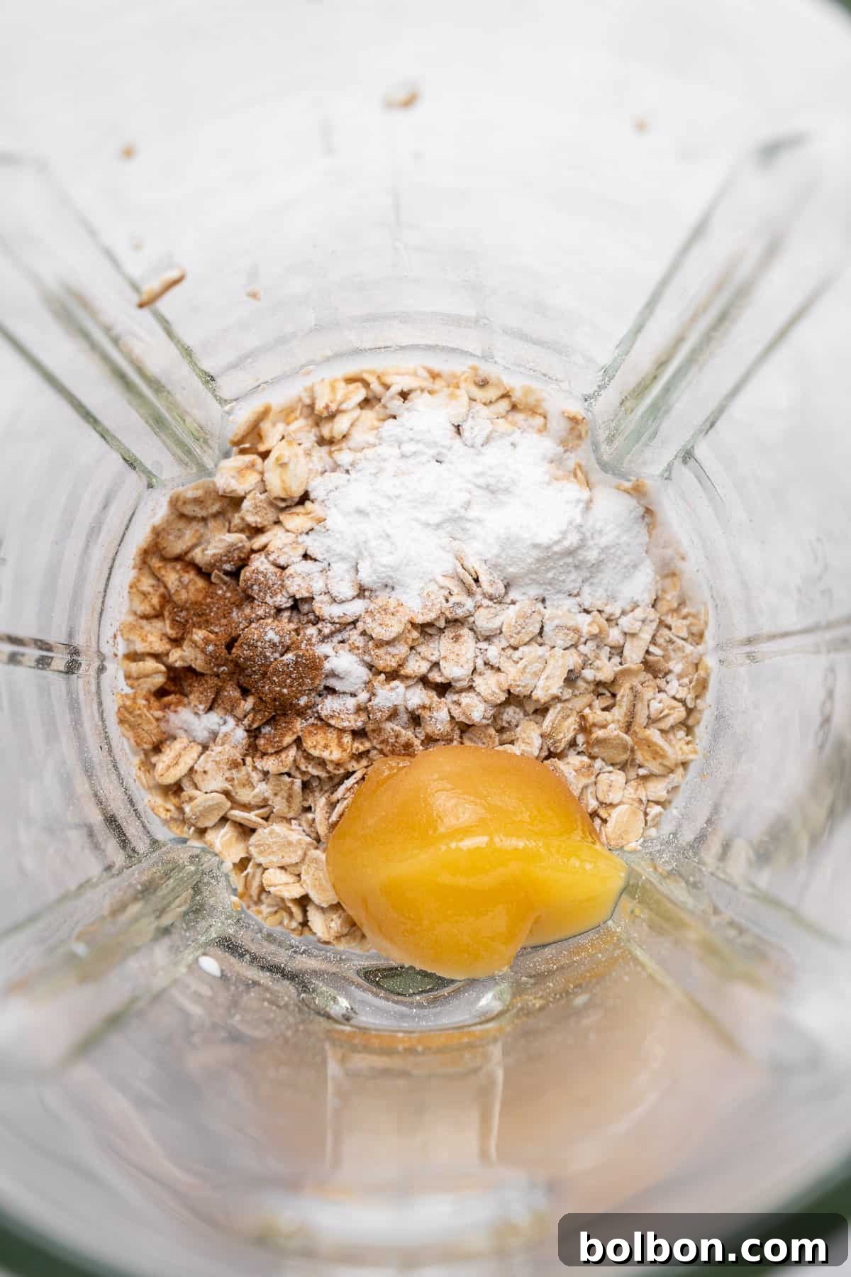 A blender contains oats, a dollop of honey, cinnamon, and a scoop of white powder, likely baking powder, seen from above. The ingredients are ready for blending, suggesting preparation for a smoothie or baking recipe.