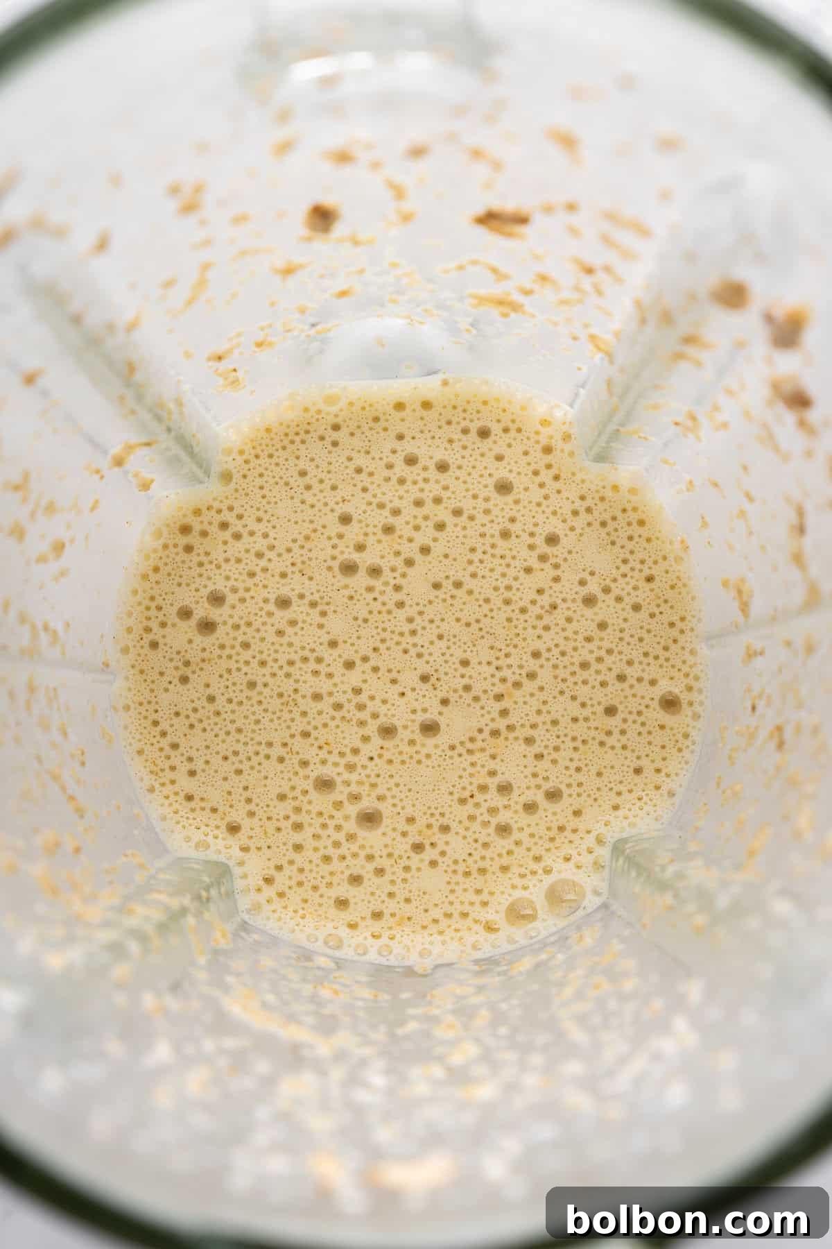 A top view of a blender with a creamy, frothy beige liquid inside, likely a smoothie or shake. The blender shows some light residue on the sides, suggesting recent blending.