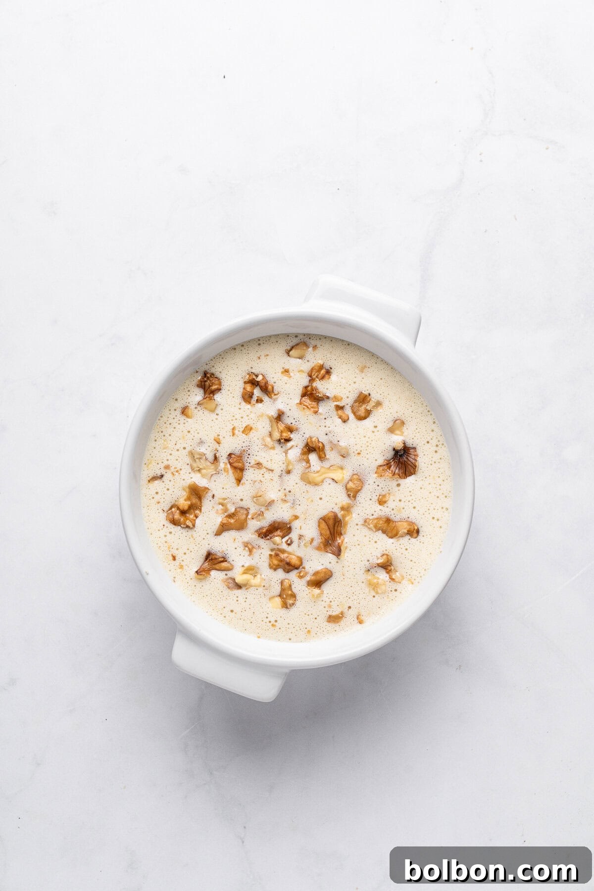 A round white dish filled with creamy pudding, topped with chopped walnuts, sits on a light gray marble surface. The pudding has a smooth texture, and the nuts add a crunchy garnish.