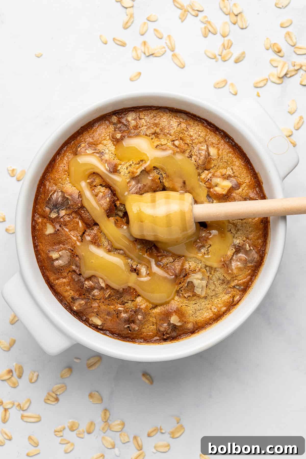 A round baked oatmeal dish in a white ceramic container drizzled with honey, shown on a light surface. Oats and a wooden honey dipper are nearby. The oatmeal has a golden-brown top with visible nuts.