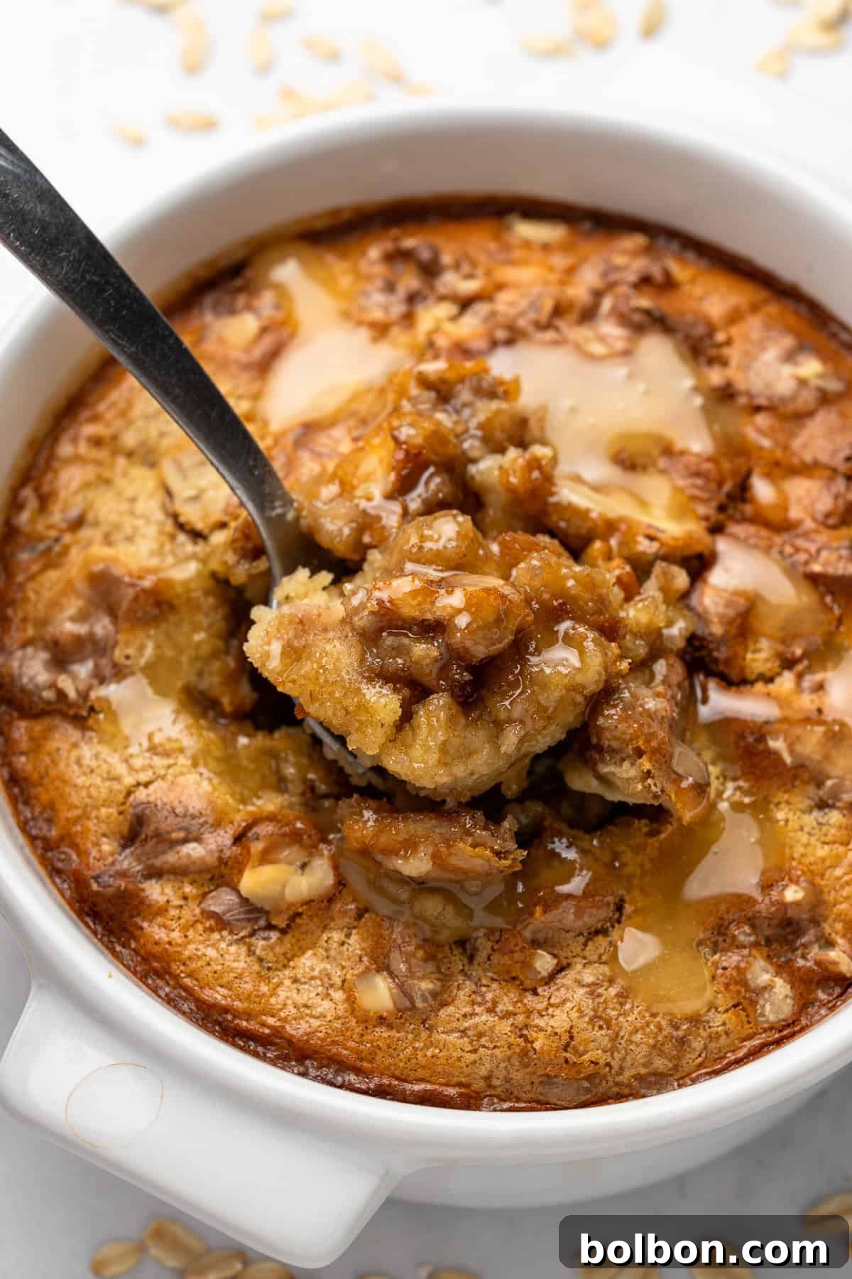 A close-up of a freshly baked dish of apple pie oatmeal in a white bowl. The oatmeal is topped with baked apple pieces and drizzled with syrup. A spoon is scooping a portion from the middle, and a few oats are scattered around the bowl.