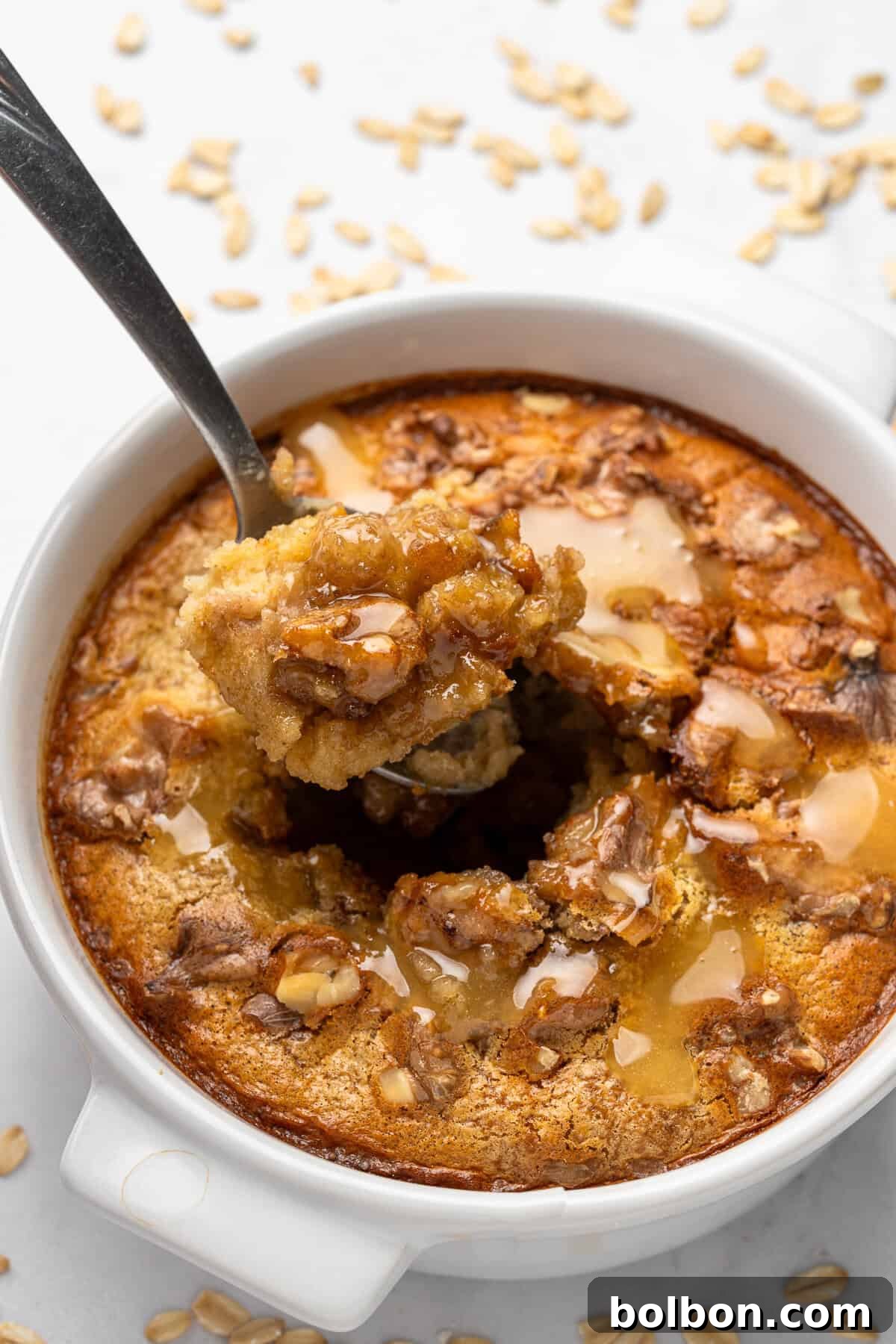A spoon scoops a portion of freshly baked oatmeal dessert from a white ceramic dish. The dish is topped with a drizzle of honey and sprinkled oats. Flecks of brown sugar or nuts add texture to the golden-brown surface.