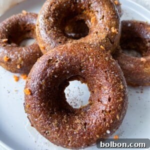 Four Pumpkin Spice Donuts with a brown, slightly glossy surface are stacked on a white plate. Small orange flecks peek through the donuts, and a white cloth is partially visible in the background.