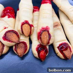 A close-up of a perfectly shaped Witch Finger Cookie with a blanched almond nail and a touch of cherry jam, showcasing its buttery texture and spooky detail.