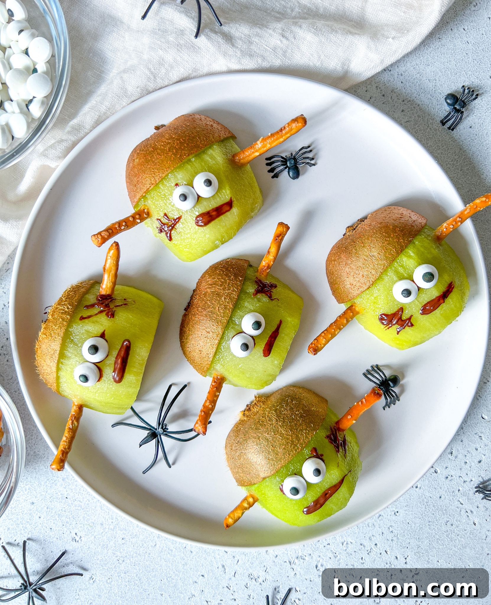 Spooky Franken-Kiwis 2 Two vibrant green kiwis, expertly transformed into adorable Frankenstein monsters, stand upright. Their faces are decorated with white candy eyes, intricate chocolate mouths, and playful 'scars.' The natural kiwi skin is ingeniously used to create a jagged 'hairline,' adding to their charm. These wholesome and fun treats are presented on a rustic wooden board, perfectly embodying the spirit of a healthy and creative Halloween snack. The image captures the inviting blend of spooky and cute, making them an ideal festive addition.