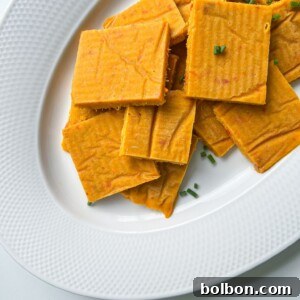 Rectangular, orange-colored crackers or chips stacked on a white textured plate, with a few pieces garnished with small green herbs—these snacks are inspired by the savory flavor of Vegetable Stock Cubes.