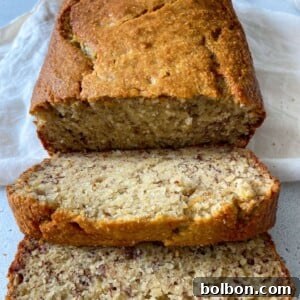 A loaf of Gluten Free Banana Bread with a golden-brown crust, partially sliced to reveal its moist, speckled interior. Two slices are laid in front on a white surface with a white cloth underneath.