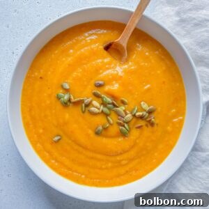 A bowl of creamy Roasted Butternut Squash Soup topped with pumpkin seeds, with a wooden spoon resting inside. The bowl sits on a light surface next to a textured white cloth.