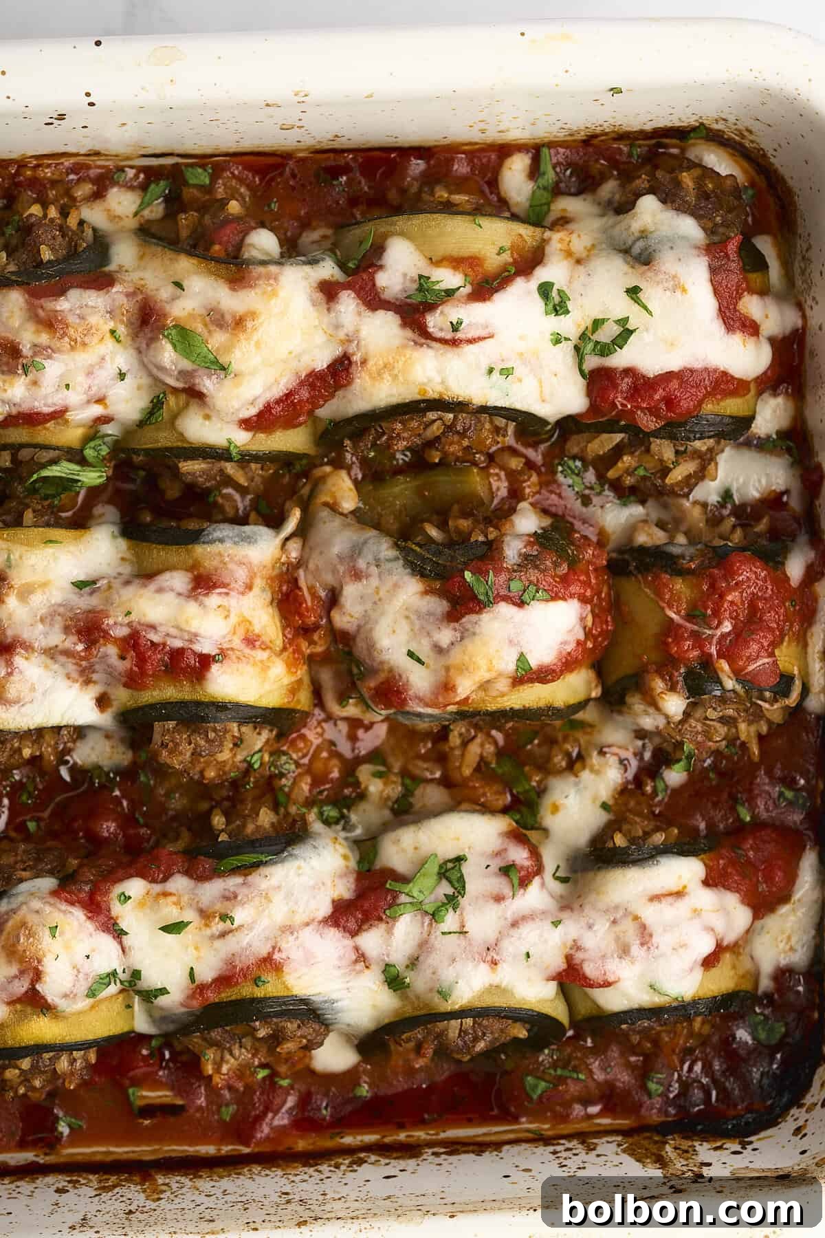A close-up of a baked dish featuring zucchini boats stuffed with meat and topped with melted cheese and tomato sauce, garnished with chopped parsley.