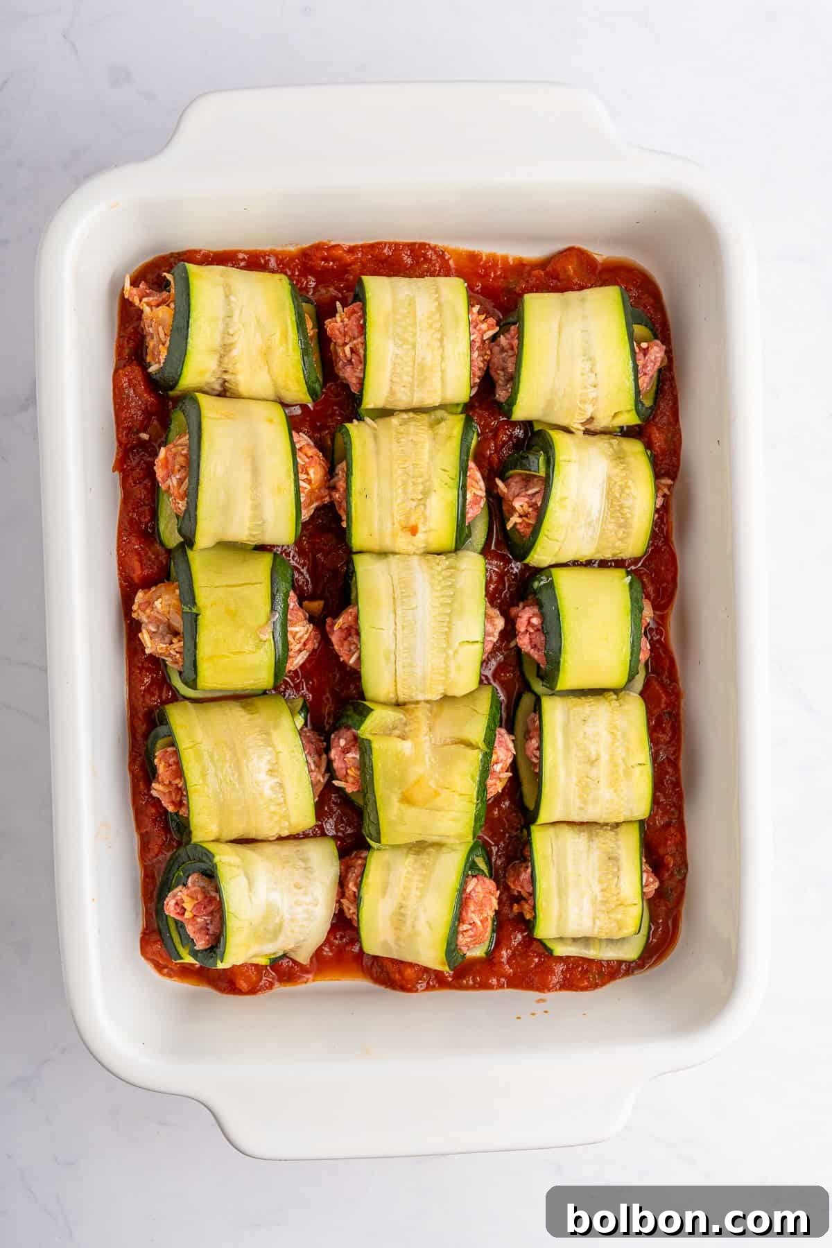 A white baking dish filled with zucchini roll-ups on a bed of tomato sauce. The roll-ups are made with thin zucchini slices wrapped around a filling, possibly meat or a plant-based alternative. The dish sits on a white marble surface.