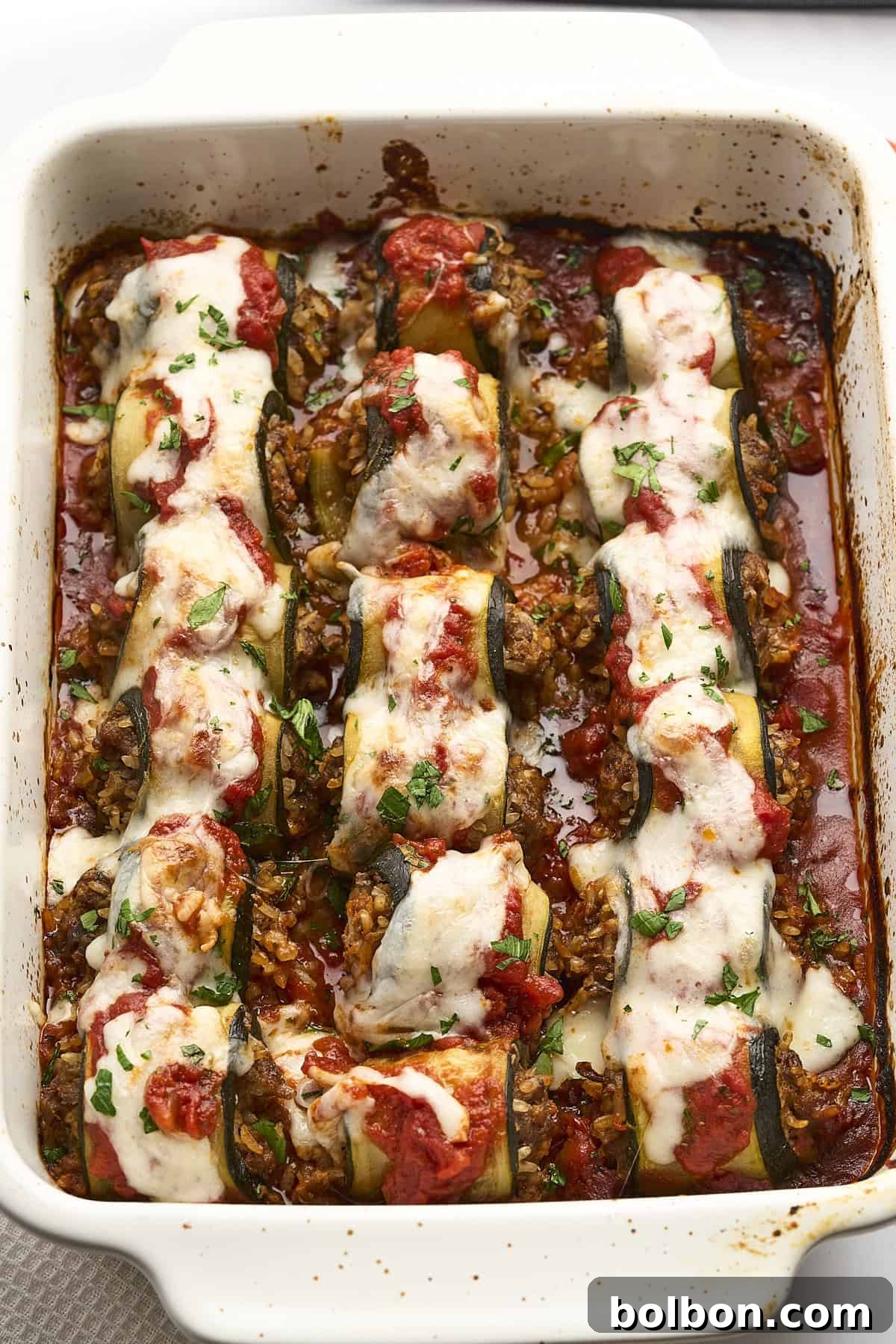 A baked dish featuring zucchini roll-ups filled with a savory mixture, topped with melted cheese and tomato sauce. The roll-ups are garnished with chopped herbs, all within a rectangular baking dish.