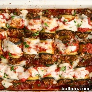 A baked dish of rolled zucchini slices filled with a savory mixture, topped with tomato sauce, melted cheese, and fresh herbs, all in a white rectangular baking dish. The dish appears freshly cooked and garnished.