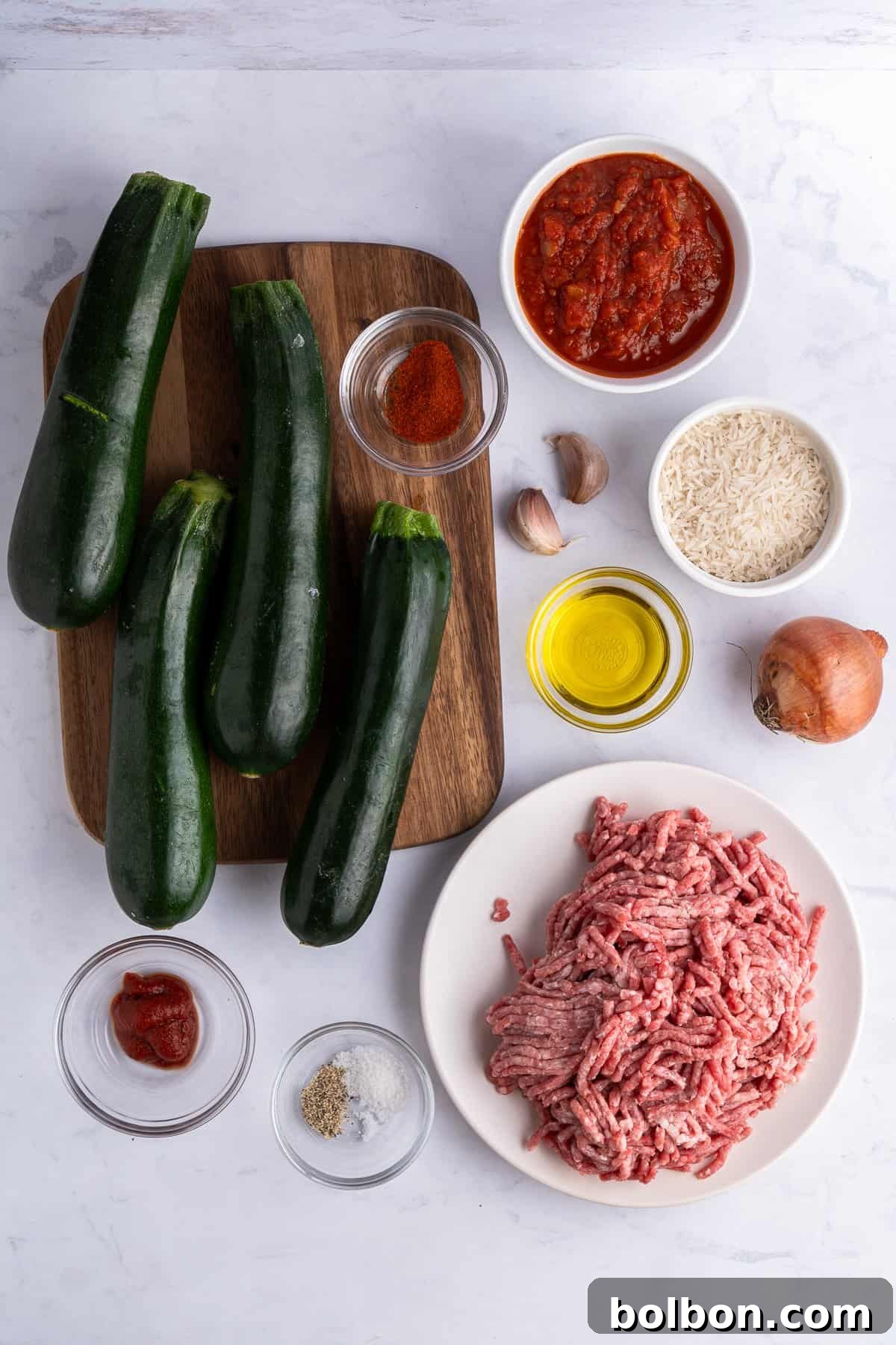 Ingredients for a dish arranged on a marble surface: several zucchinis on a wooden board, bowls of tomato sauce, rice, ground meat, spices, olive oil, and small bowls containing tomato paste, salt, pepper, garlic, and a whole onion.