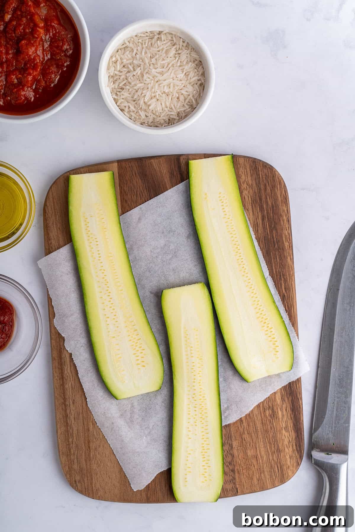 Sliced zucchini on a wooden board with parchment paper, surrounded by bowls of rice, tomato sauce, and olive oil. A knife is visible on the right side.