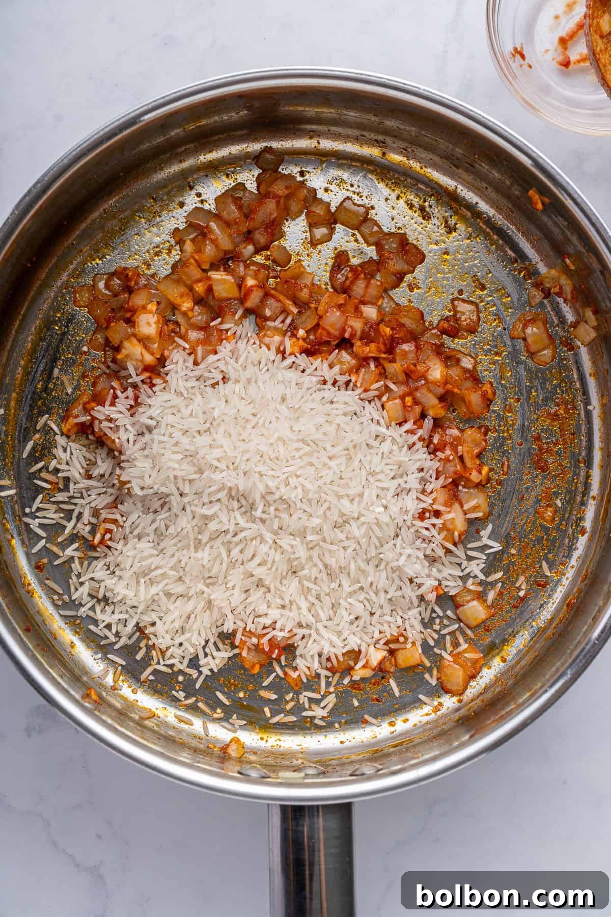 A stainless steel pan on a marble surface contains sautéed onions and a heap of uncooked white rice. The onions appear to be seasoned with spices, giving them a reddish-brown color.