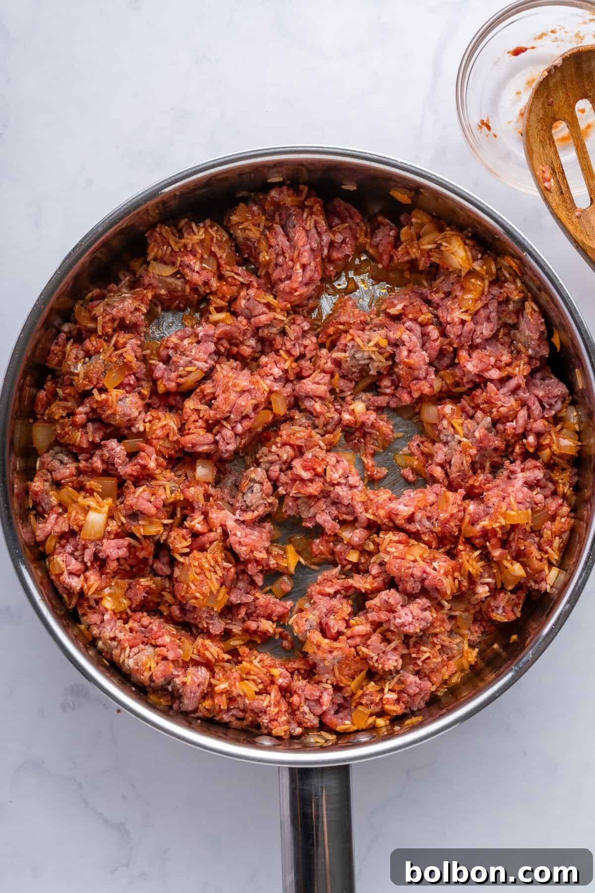 A frying pan filled with minced meat cooking on a stovetop. The meat is partially cooked, mixed with chopped onions and other ingredients, and has a reddish hue. The background is a light, textured surface. A wooden spoon rests nearby.