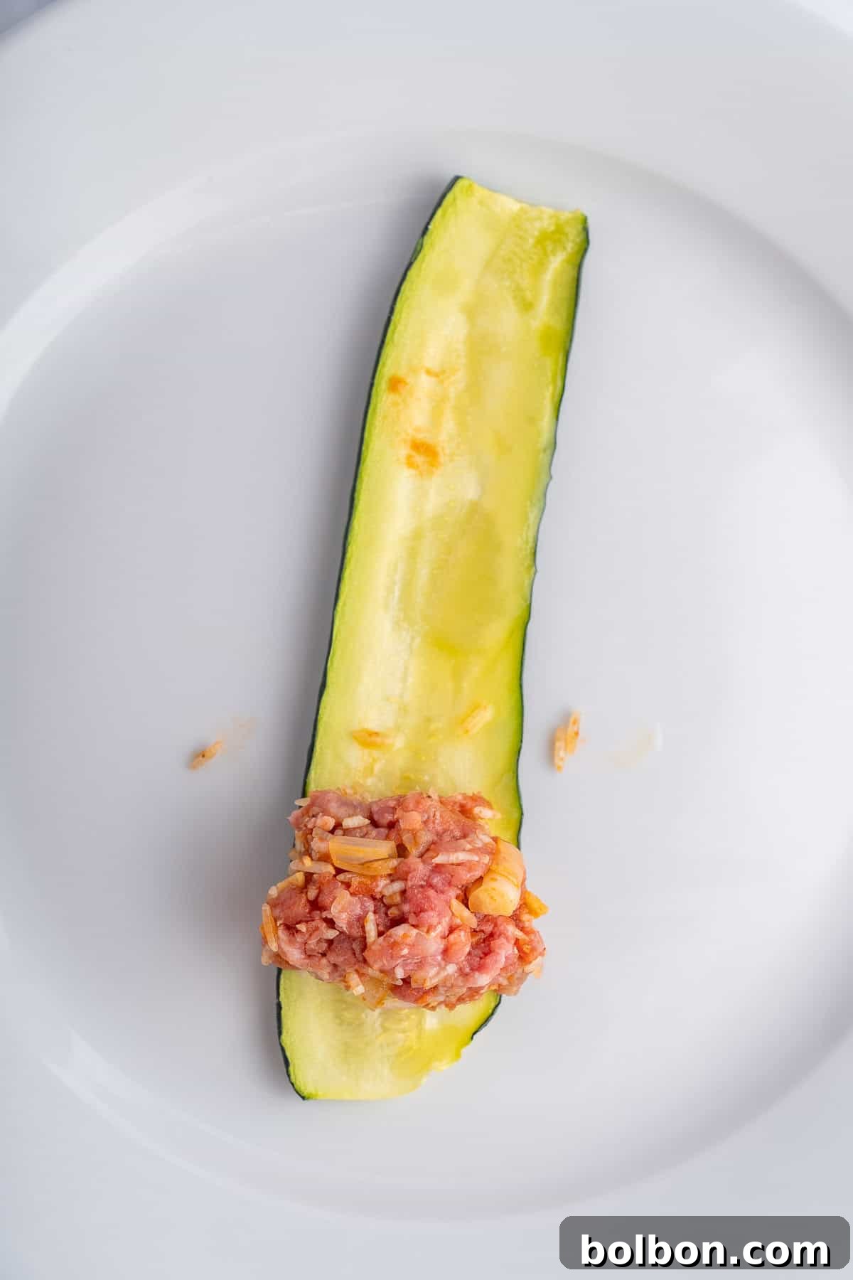 A single slice of zucchini on a white plate, hollowed out and topped with a small portion of minced meat mixed with onions.