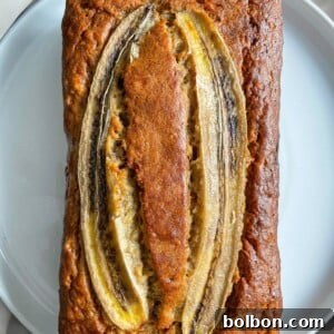 A loaf of banana bread topped with three lengthwise slices of banana sits on a white plate, showcasing its golden-brown crust and moist texture.