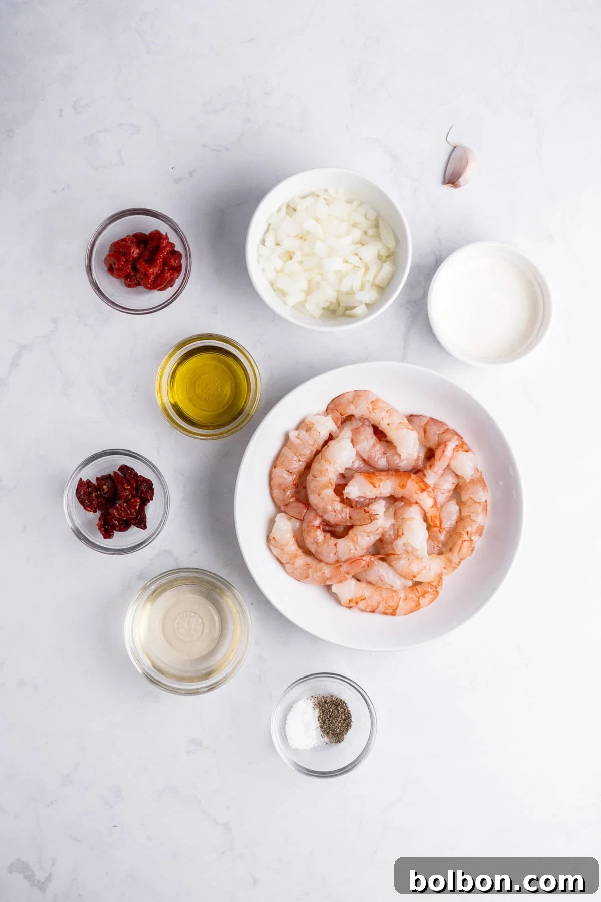A flat lay of shrimp in a white bowl surrounded by bowls containing chopped onions, tomato paste, sun-dried tomatoes, olive oil, cream, white wine, a garlic clove, and a small dish of salt and pepper on a light marble surface.