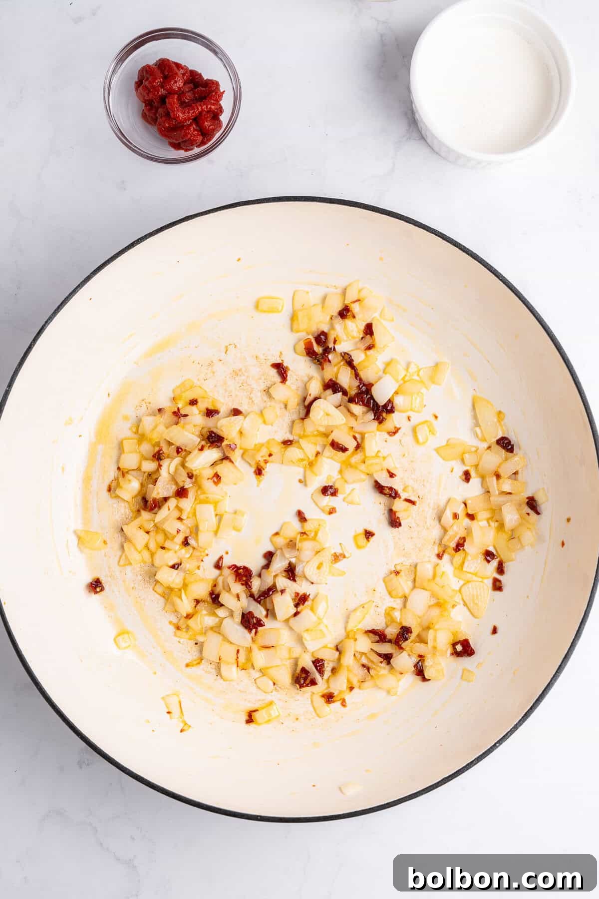 A white skillet on a marble surface contains sautéed onions and spices. Nearby are small bowls with tomato paste and cream.