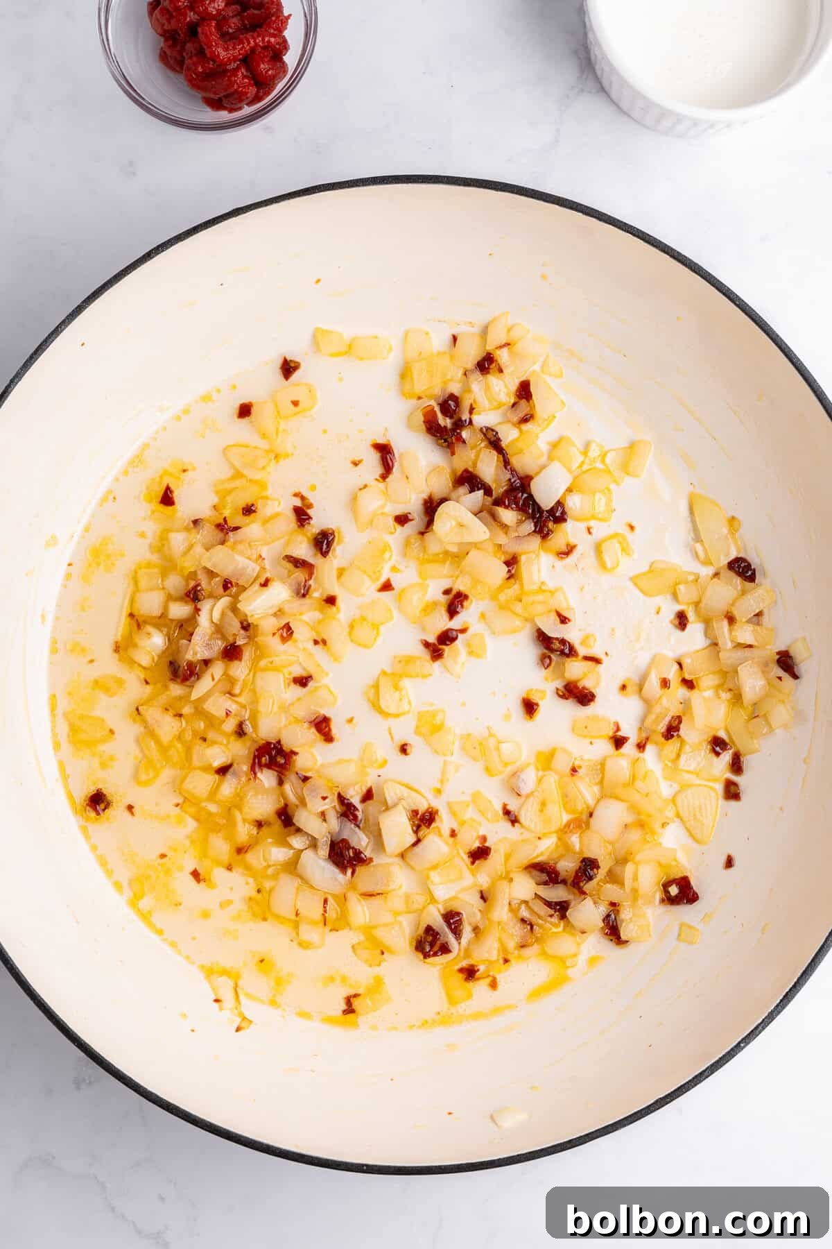 Chopped onions and red pepper flakes sautéing in oil in a large white sauté pan. Bowls with ingredients, including tomato paste and cream, are placed nearby on a white marble surface.