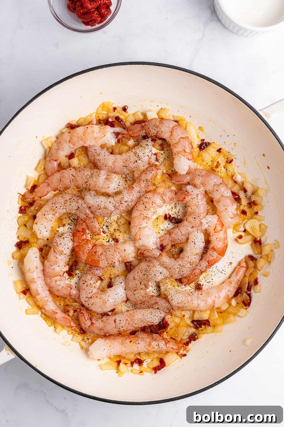 A pan filled with raw shrimp seasoned with pepper, resting on sautéed onions and diced sun-dried tomatoes. A small bowl of tomato paste is visible in the top left corner. The background is a white marble surface.