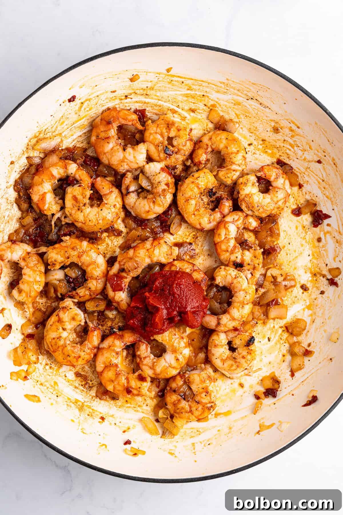 A white pan filled with cooked shrimp, chopped onions, and a blend of spices. A dollop of tomato paste is in the center, and everything is mixed together with visible seasonings. The background is a light, smooth surface.