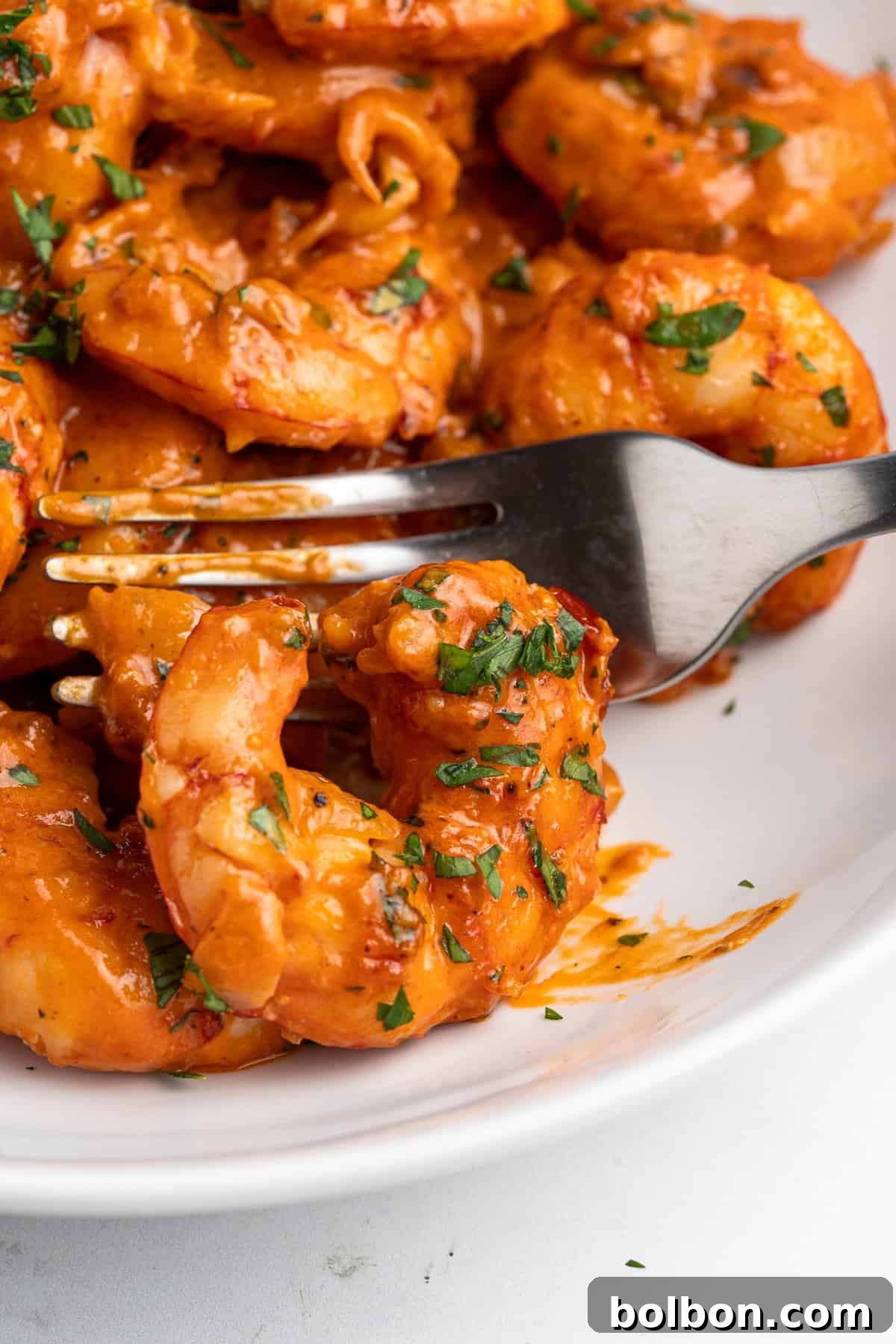 Close-up of a fork holding a creamy tuscan shrimp coated in a rich, creamy sauce with finely chopped herbs on a white plate. Other shrimp are visible in the background, all garnished with herbs.