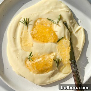 A white plate with creamy yogurt and whipped Brie, topped with honeycomb pieces and rosemary sprigs; a butter knife rests on the plate.