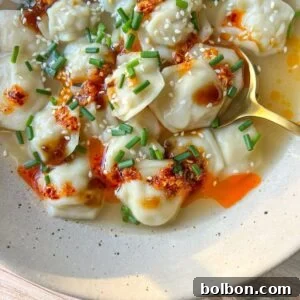 A bowl of Dumpling Soup in broth, topped with chili oil, sesame seeds, chopped chives, and drizzled sauce, served with a golden spoon on the side.