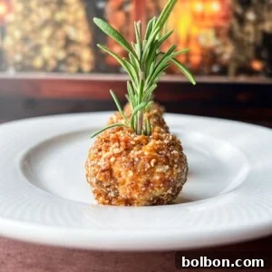 A close-up of Holiday Cheese Balls, a round, breaded appetizer garnished with a fresh sprig of rosemary.