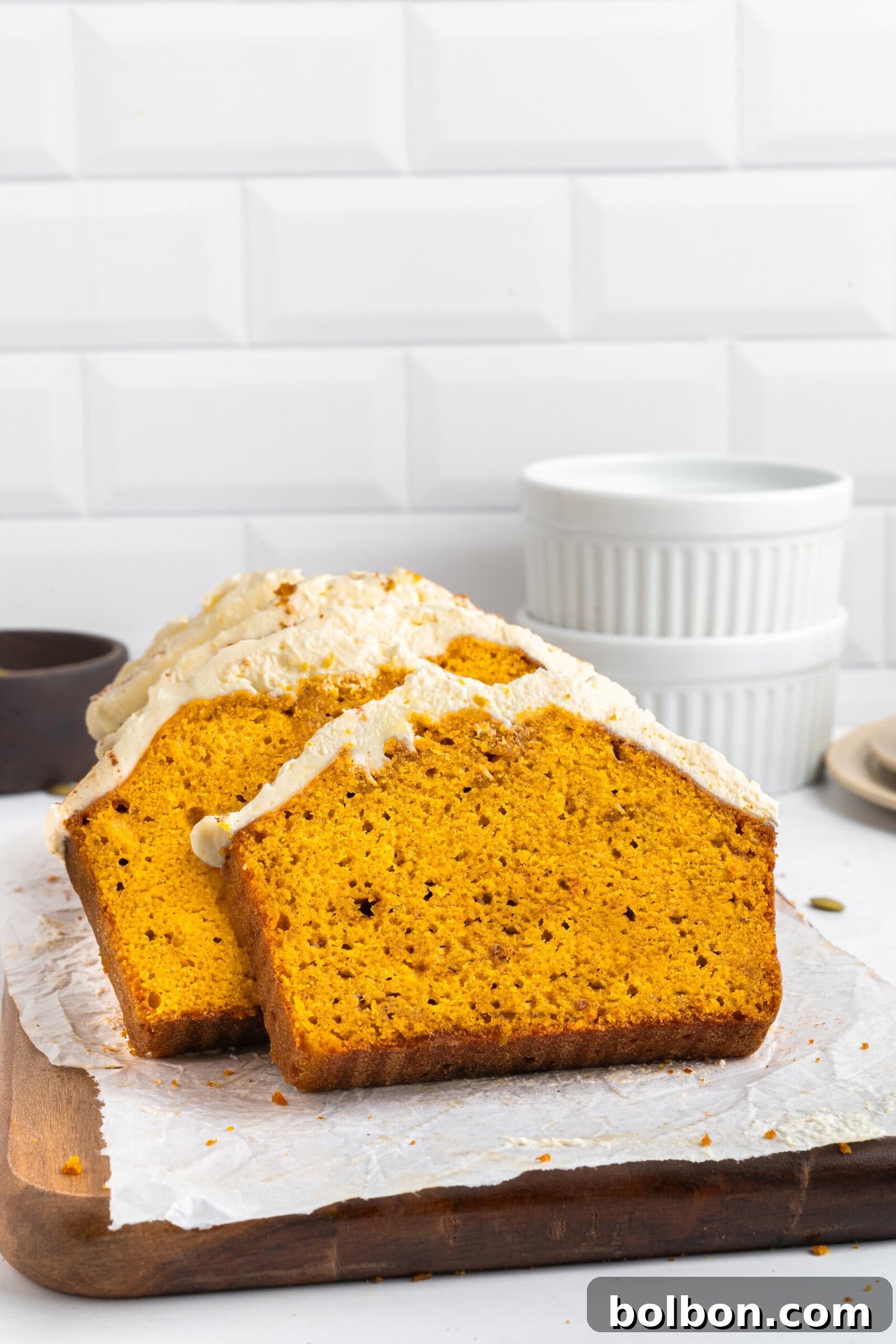 Two thick slices of bright orange pumpkin bread with cream cheese frosting sit on parchment paper atop a wooden board, with stacked white ramekins and a white tiled wall in the background.
