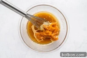 A metal whisk mixing orange pumpkin puree with brown liquid ingredients in a clear glass bowl on a white countertop.