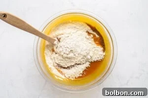 A glass bowl containing a mixture of flour and a yellow batter sits on a white surface. A wooden spoon rests inside the bowl, partially submerged in the mixture, suggesting the ingredients are being combined.