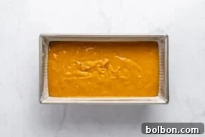 A rectangular metal baking pan filled with smooth, orange-yellow batter sits on a white marble surface, ready to be baked.