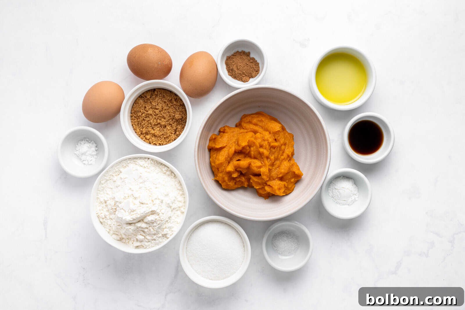 Bowls containing flour, sugar, brown sugar, baking powder, spices, salt, and vanilla surround a bowl of pumpkin puree. Three eggs and a small bowl of oil are also included, all arranged on a white surface.
