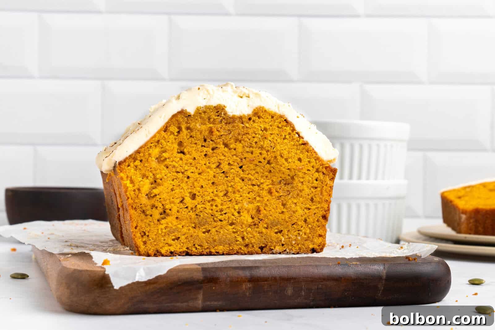 A loaf of pumpkin bread with white frosting sits on parchment paper atop a wooden cutting board, with a slice cut off and dishes in the background.