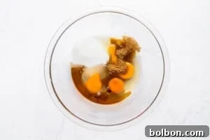 A glass bowl containing cracked eggs, white sugar, brown sugar, and vanilla extract sits on a white countertop, viewed from above.